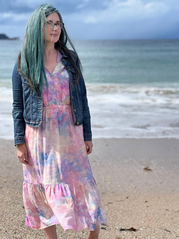 this is willow founder wearing the pink ecovero quinn midi-dress with a denim jacket. Lynda is on a beach in Northern Ireland, with the sea behind her. The wind is blowing her midi-dress forward.