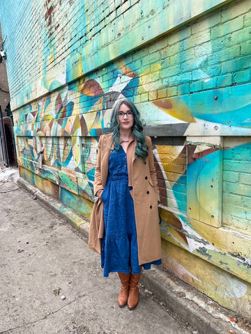 this is willow founder Lynda is wearing the Quinn midi-dress in deadstock linen. Lynda is standing in front of some street art, wearing a long sweater coat in camel, a long camel coat, and caramel riding boots