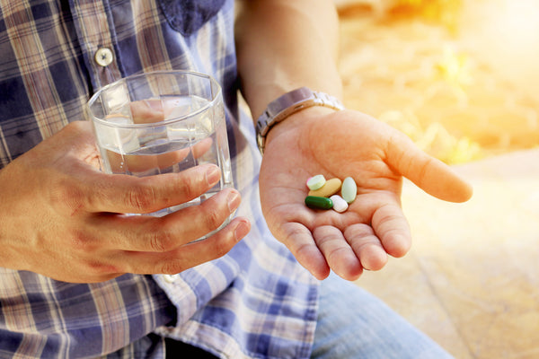 various supplement tablets and capsules in a man's palm with him holding a glass of water in the other hand