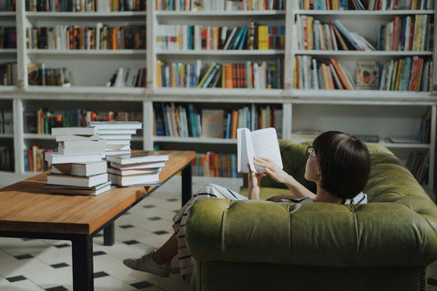 Sit back, read, and relax at the library for some quality “me time” of your own. Photo by Cottonbro Studios. A woman reading on a green sofa, next to a table with a pile of books on top of it.