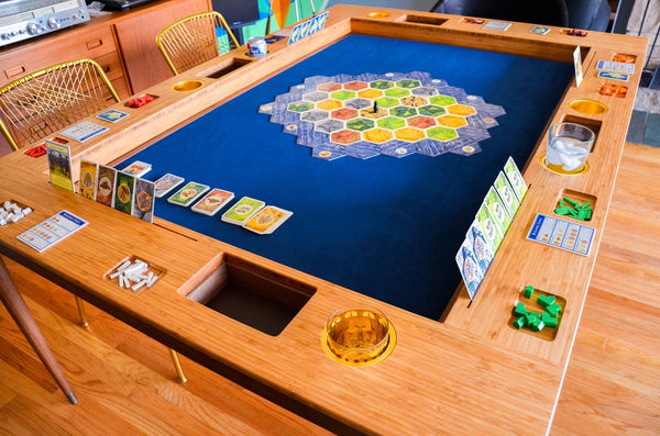 The same mid century modern table as above with the top removed. Inset into the table is a blue game mat with teh game Settlers of Catan being played. Along the edges of the table are cup holders and carved holders for game pieces.
