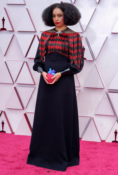 Celeste Wait wearing a Gucci gown. the top bodice features a red and black harlequin pattern and the skirt is a black a-line. She's holding a clutch that looks like a human heart decorated with crystals. 