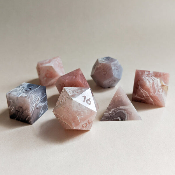 A set of handcrafted resin tabletop rpg dice ranging from D20 to a D4. The colors are pink and grey marble with unpainted numbers. Created by Yaniir