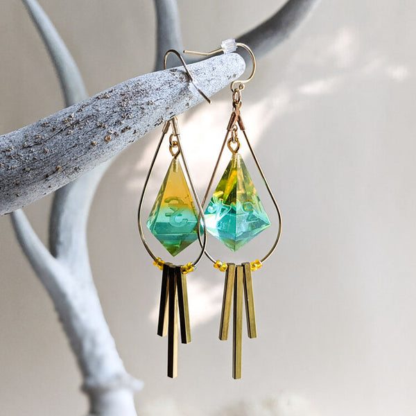 A set of earrings created with resin D8 dice. The dice are aqua and apple green surrounded by a gold metal wire and gold metal bars. Created by Yaniir