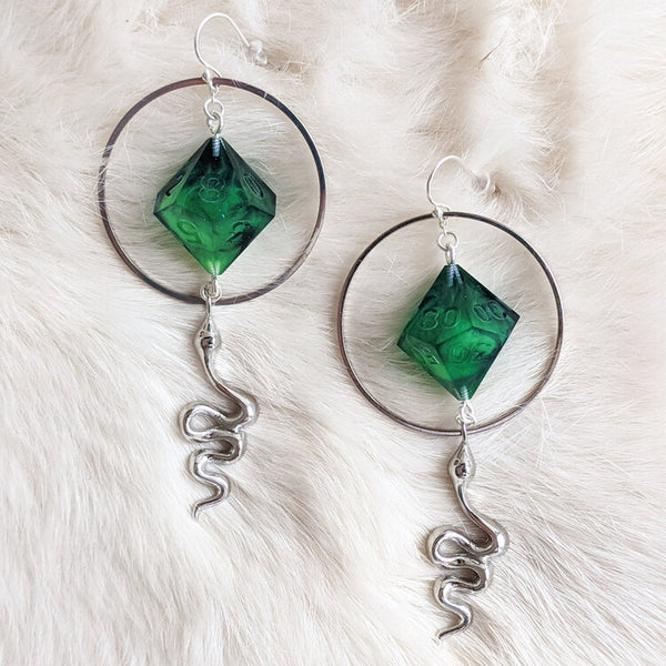 A set of earrings created with resin D10 dice. The dice are translucent green surrounded by a silver metal wire with silver snakes hanging from it. Created by Yaniir