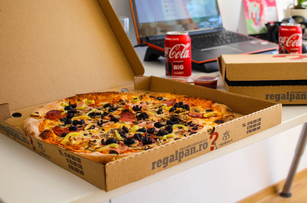 Boxes of pizza on a desk. One box of pizza is open to reveal a pizza, the other one is partially closed. Two cans of coke are also on the desk.