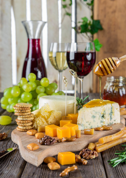 A charcuterie board covered in a variety of cheese, crackers and nuts. A bunch of grapes and wines are behind the board. A honey dipper is drizzling honey on top of one of the cheeses.