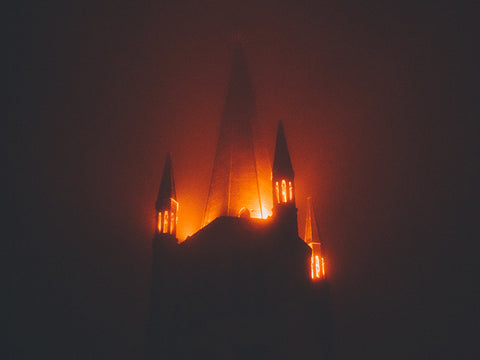 The silhouette of a castle against a dark background. at the top of the castle an orange light glows as if it's on fire.