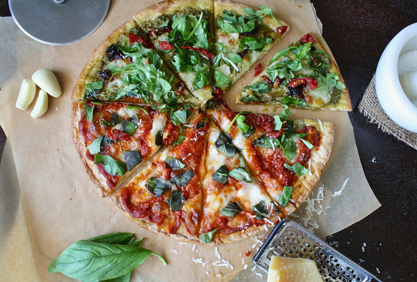 A top down view of a handmade pizza. Half of the pizza has a pesto sauce nd various toppings. The other half has red sauce, cheese, and basil. There's garlic, parmesan cheese, and basic on the table around the pizza.