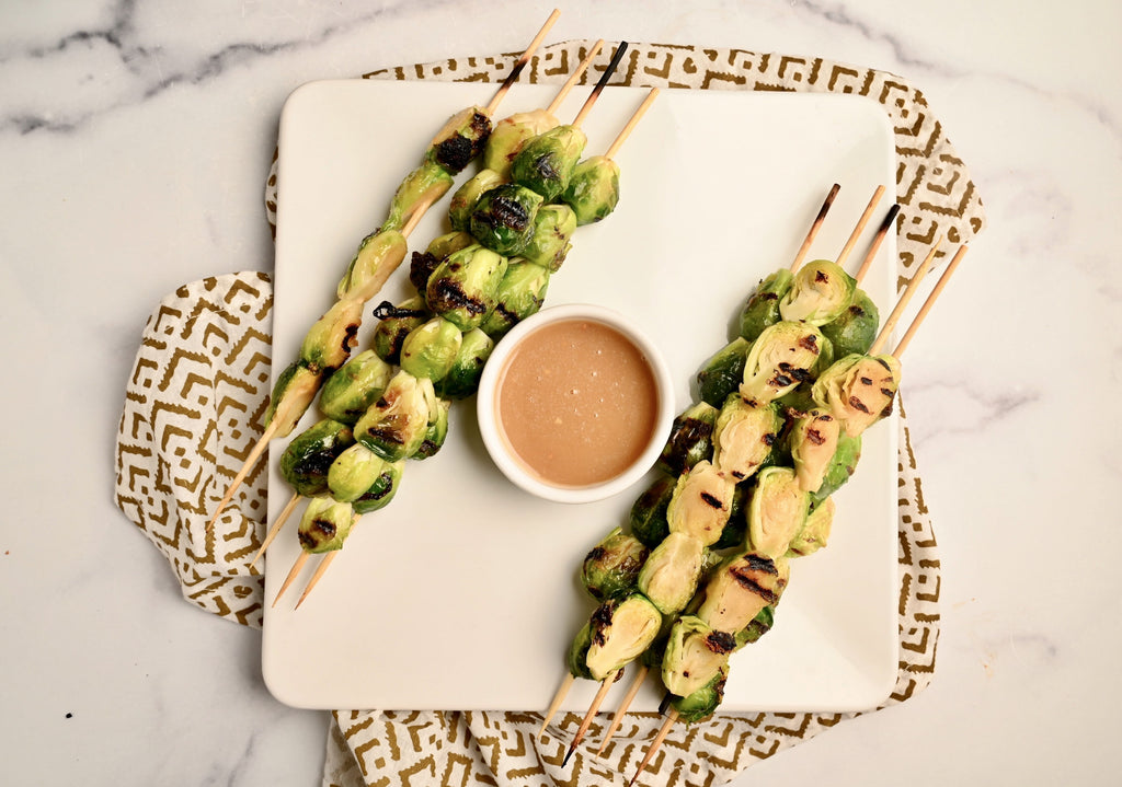 A plate of grilled Brussels sprouts on skewers next to a bowl of creamy homemade peanut sauce