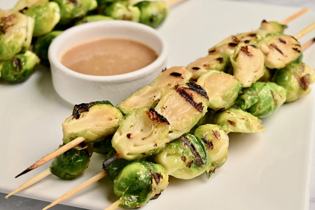 A close up of grilled Brussels sprouts on skewers next to a bowl of creamy homemade peanut sauce