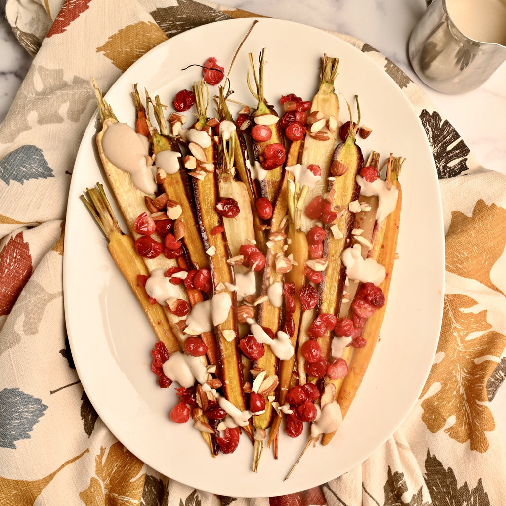 A white platter of roasted carrots with bright red cranberries on top with a drizzle of homemade nut buttersauce