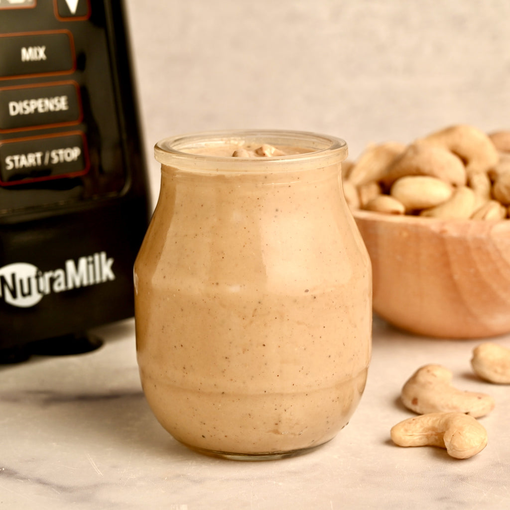 A clear glass jar filled with cashew butter with The NutraMilk off to the side