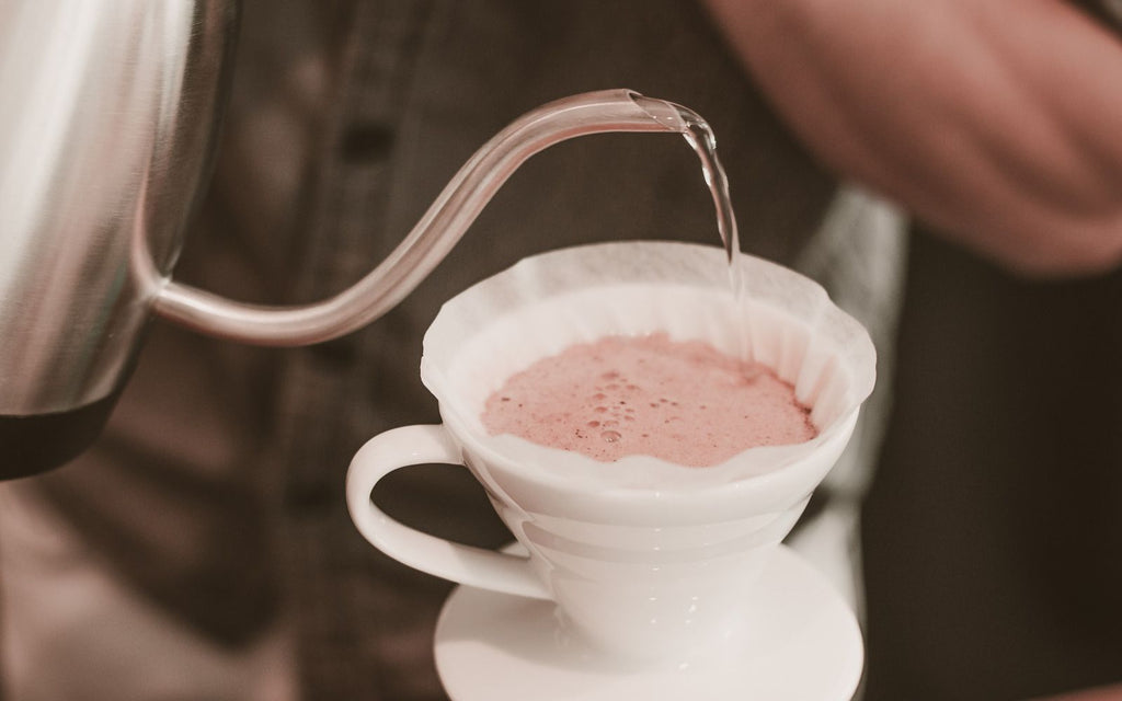 A closeup of the gooseneck spout of a kettle pouring water over coffee grounds.