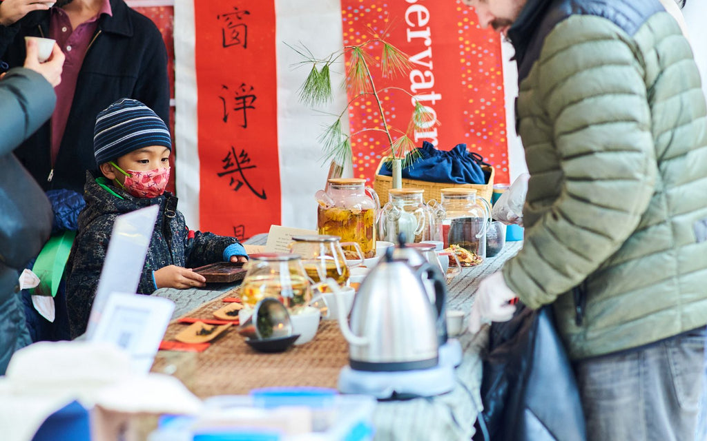 A child in a mask is being served tea at an outdoor event.