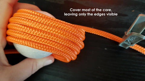 Multiple loops of orange rope are wrapped around a ball in the first layer of a monkey fist