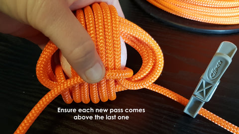 Multiple loops of orange rope are wrapped around a ball, working on the third layer to make a monkey fist