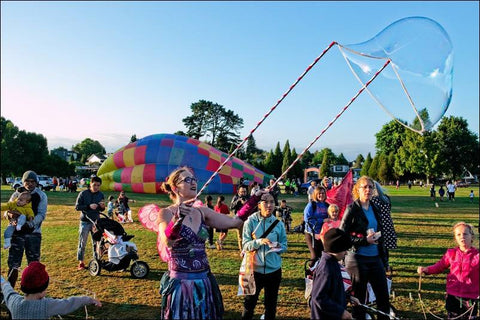 A 'fairy' holds two sticks with a giant bubble forming while children watch 