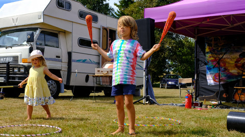 A young boy spins sock poi while a young girl plays with a spin staff behind him, and there are hula hoops on the ground near them