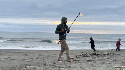 A man spins fire spin staff on the beach while children play in the background