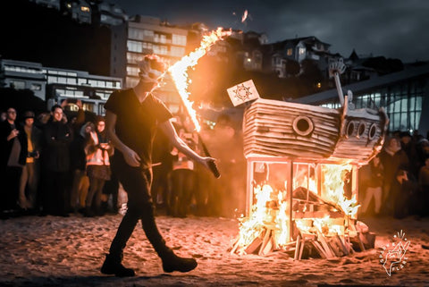 A person in a mask spins a fire sword on the beach with a bonfire next to them and an audience in the background