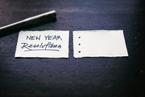 Two small pieces of paper sit on a black desk with a pen, one paper says 'new year resolutions'