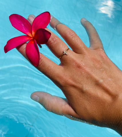 Hand mit funkelndem Kreuz Ring im Pool Wasser mit rosa roter Blume. Der Kreuz Ring ist mit Zirkonen besetzt und soll das wasserfeste Material Stainless Steel zum Ausdruck bringen.