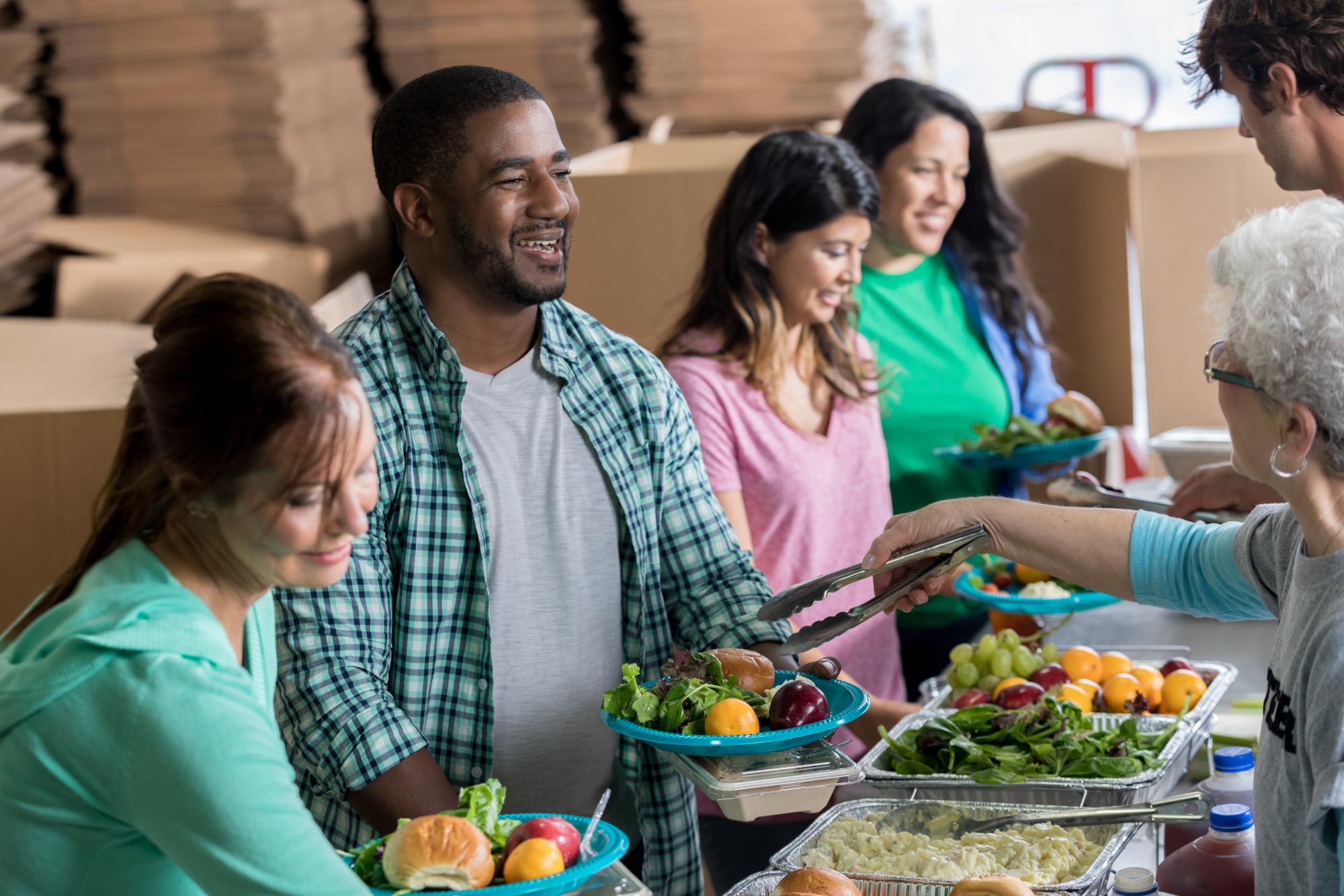 Diverse people receive a healthy meal in a local soup kitchen. A senior woman is serving fruit and salad to a mid adult man. Trays of healthy food are on the table.