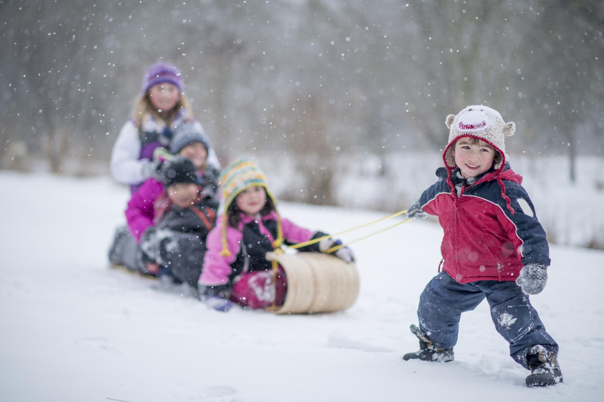 A little boy is trying to pull a sled filled with children. They are outside playing in the snow and wearing winter clothes.