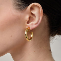 A model wearing the Allie Hoop Clip On Earrings in Gold, which are perfect for all ear types. The Mosquito Coil Clip-On Closure is suitable for Thick/Large, Sensitive, Small/Thin, Stretched/Healing, and Keloid Prone Ears. Made of a Gold tone copper alloy, each purchase includes one pair.