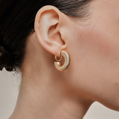 A model wearing the Bold Ribbed Hoop Clip On Earrings in Gold. Our screwback closure and manual adjustment capabilities cater to a wide range of needs, including thick, sensitive, small, and keloid-prone ears. Easily wear them for 8-12 hours for all-day fashion. Sold in pairs.