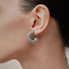 A model wearing the Bold Ribbed Hoop Clip On Earrings in Silver. Our screwback closure and manual adjustment capabilities cater to a wide range of needs, including thick, sensitive, small, and keloid-prone ears. Easily wear them for 8-12 hours for all-day fashion. Sold in pairs.