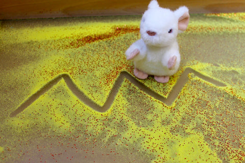 A zigzag pattern has been drawn in yellow sand. Next to it sits a white toy mouse.