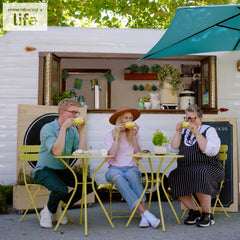 Photo Credit: Alex Rhoades, from "The Gentle Art of Swedish Death Cleaning" Photo Description: The co-hosts of "The Gentle Art of Swedish Death Cleaning," Johan Svenson, Katarina Blöm and Ellinor Engström, are seated at yellow cafe tables in front of a coffee trailer, sipping coffee.