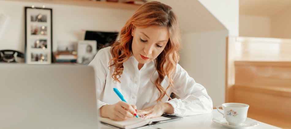 Image of woman writing in a notebook. There is a cup of coffee to one side of her and a laptop on the other. Photos of people are on the wall behind her.