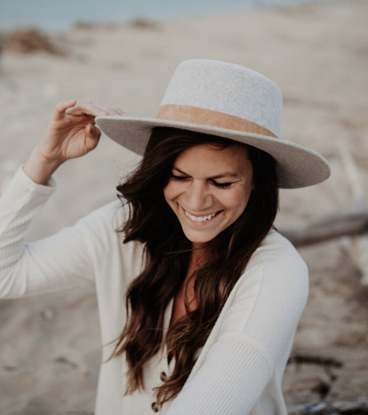 Portrait of Dana Frost. She is on the beach, wearing white and a white hat. Her eyes are cast downward. She has long brown hair and is smiling.