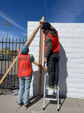 Two artists in orange safety vests use a large wooden compass to mark angles on a white wall.