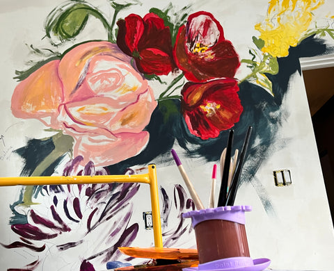 A mural in progress. The foreground is yellow scaffolding holding a purple rinse cup with multiple brush handles sticking out of it. The background shows a mural of a floral bouquet sketched on a white wall, with color tests of yellow, peach, navy, purple, and various red tones showing where the flowers will bloom
