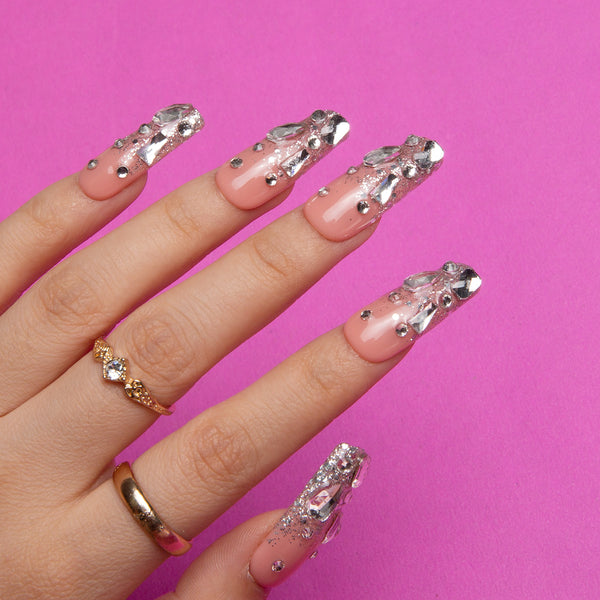 Hand wearing long coffin-shaped press-on nails with silver glitter and rhinestones against a pink background. Features gold rings on the fingers.