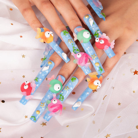 Long, stiletto-shaped acrylic nails painted a light blue with 3D fish charms, glitter, and small seashell accents.