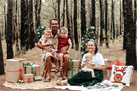 Family Christmas photo in pine forest. Father sitting on chair with one year old and three year old daughter on knees. Mother sitting on rug on the ground with two month old baby boy. Christmas presents and cushions layed out on the rug and around chair.