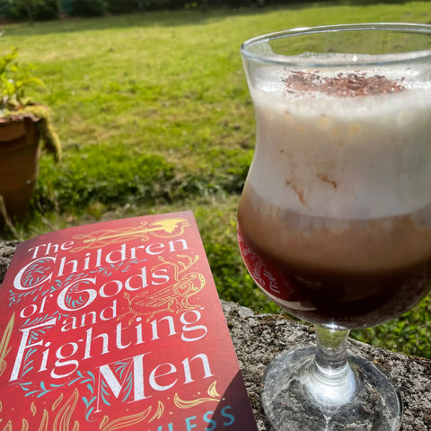 A photograph of a sunny garden. Resting on the garden wall is a paperback of The Children of Gods and Fighting Me by Shauna Lawless, an Irish author, along with a tall glass of Maple Iced Coffee. The image is intended to convey the idea of relaxing on a sunny evening with a good book and a cold refreshing drink.