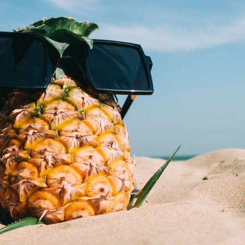A picture of a pineapple on a sandy beach. The pineapple is wearing sunglasses.