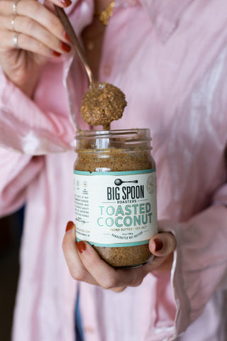A person pulls Toasted Coconut Almond Butter out of a jar. They are wearing pink.
