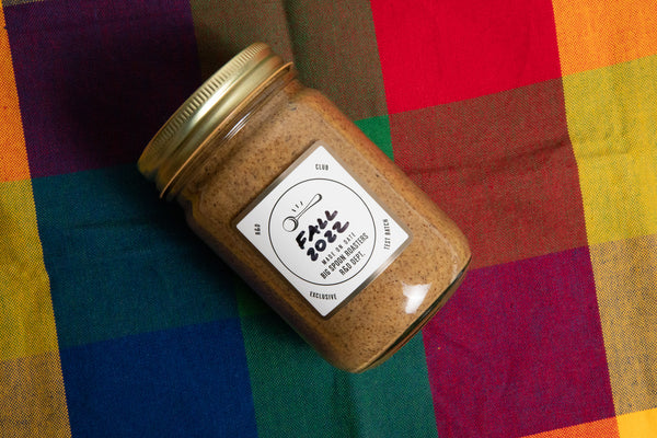 A jar of our '22 Q4 R&D Batch, Hummingbird Cake Almond Butter, sits atop a multicolored plaid background. 