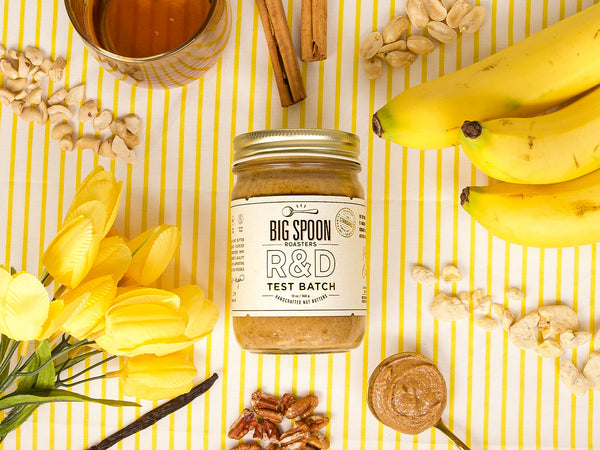 13oz jar jar of R&D Bananas Foster Peanut & Cashew Butter surrounded by bananas, yellow tulips, peanuts, cashews, rum, cinnamon sticks, and banana chips with a yellow-striped background
