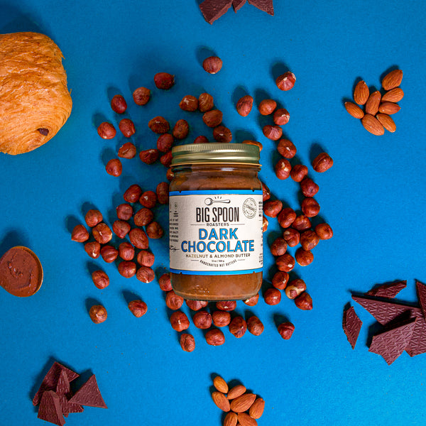 13oz jar of Dark Chocolate Hazelnut & Almond Butter surrounded by hazelnuts, chocolate, almonds, a spoon of nut butter, and a dark chocolate croissant with a rich blue background.