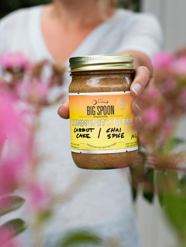 A person holds a jar of Community Blend nut butter. They are near pink flowers.