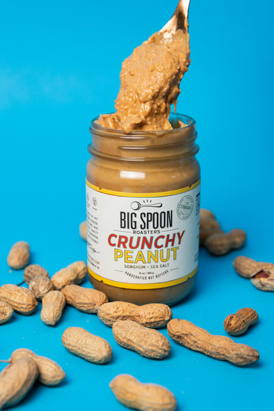 A spoon pulls crunchy peanut butter out of a Big Spoon Roasters Crunchy Peanut jar. The background is bright blue and there are shelled peanuts on the ground.