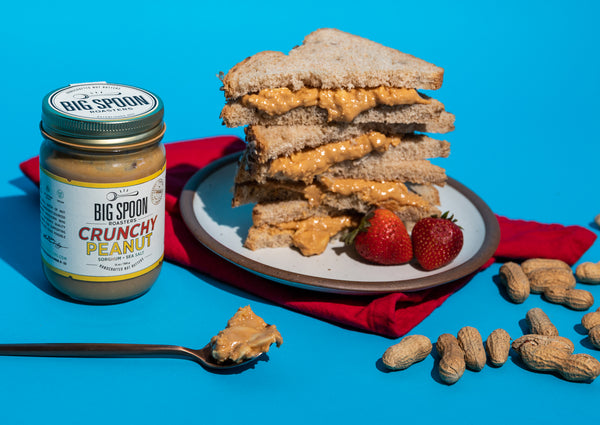 A stack of crunchy peanut butter sandwiches is piled high on a white plate against a bright blue backdrop. A jar of Crunchy Peanut is to the left, there is a spoon with crunchy peanut butter on it in the foreground, and shelled peanuts are scattered about.